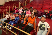 Elementary students sitting in two rows of red theatre seats with empty rows behind them.