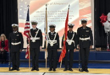 A group of six uniformed students stand in a straight line as an honor guard in a gymnasium. Two students on the ends hold rifles, while two in the center hold flags, including the U.S. flag and a red military-style flag.