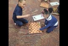Two students sitting on either side of a chess board and are mid-game.