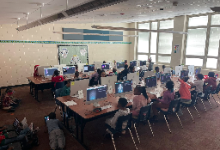 A computer lab filled with elementary students seated at rows of desktop iMac computers. Students are working individually at their stations, with monitors displaying what appears to be educational software or digital learning activities. The bright class