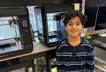 "A smiling student wearing a navy and white striped sweater stands in a classroom beside shelving units containing various supplies and equipment. Computer monitors are visible on a desk in the background."