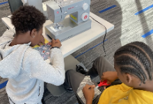 Two students work together in a Makerspace classroom. One operates a sewing machine while the other cuts fabric with scissors. Both are focused on creating their drawstring bags as part of a sewing project.
