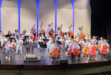 A large group of middle school musicians sits and stands on a stage holding string instruments, including violins, violas, cellos, and basses. They are wearing matching light blue shirts and smiling toward the audience, with a blue-lit backdrop behind the