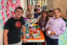 A group of elementary school students stand smiling in front of a long prize table filled with small toys, stickers, and trinkets. Colorful paper hearts with handwritten notes cover the wall behind them. Other students browse the table in the background i
