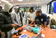 A group of high school students stand around a table as a presenter demonstrates an activity during the E4Me Career Program at the ESC of Northeast Ohio. The table is covered with materials including pamphlets, tools, and supplies, while other participant