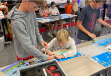 Students collaborating on a hands-on STEM project in a classroom, working with blue grid boards, small yellow and white components, and building materials spread across their desks. 