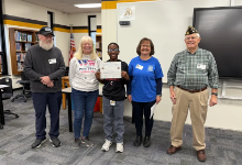 Five people stand together in a school library: four adults from VFW Post 7536 flank a student holding a certificate. 