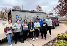 "Seven Brooklyn City Schools staff members stand in front of a Brooklyn Medical Center sign holding boxes of donated food items. The group poses outdoors on an overcast day with fall foliage visible in the background