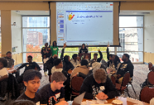 Students are sitting at round tables in their groups in front of a projection screen and appear to be participating by raising their hands. 