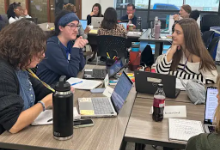 A group of educators and students sit around tables in a classroom or workshop setting, engaged in discussion and working on laptops. Papers, notebooks, and water bottles are spread across the tables as participants collaborate in small groups.