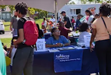 An outdoor community event showing attendees gathered around a table with a blue tablecloth bearing the "Cleveland Heights-University Heights City School District" logo. Several community members, including adults and children, are engaged in conversation