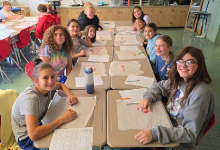  A science classroom with students seated at lab tables covered with brown paper, working on an activity. Approximately a dozen middle school students are visible, smiling at the camera while gathered around their workstations. Papers and materials are sp