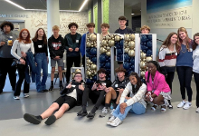 A group of nineteen Cuyahoga Heights High School students pose together in front of large balloon letters spelling "JCU" at John Carroll University after participating in the Thinkfest Ideation Workshop, with some students standing and others sitting on t