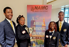 Four students in navy blazers with gold accents and khaki pants pose with a trophy beside a NAMC banner. 