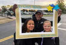 hree smiling participants at the Brooklyn My Guy Walk pose together while holding a large glittery gold picture frame decorated with blue stars and text reading 'My Guy Walk' and '#SHareYourStory'.