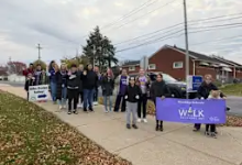 A group of approximately 15-20 people gathered on a sidewalk in front of a brick building on an overcast day. Two people in the front hold a purple banner that reads "WEEK." T
