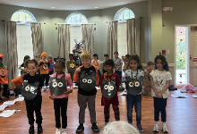 A diverse group of elementary school children stand in a line in a community room with hardwood floors and arched windows, each holding handmade black owl crafts with large white paper eyes. 