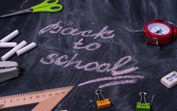 Picture of chalkboard with "Back to School" written in pink chalk, with school supplies spread on top of it, including scissors, ruler, chalk, and binder clips