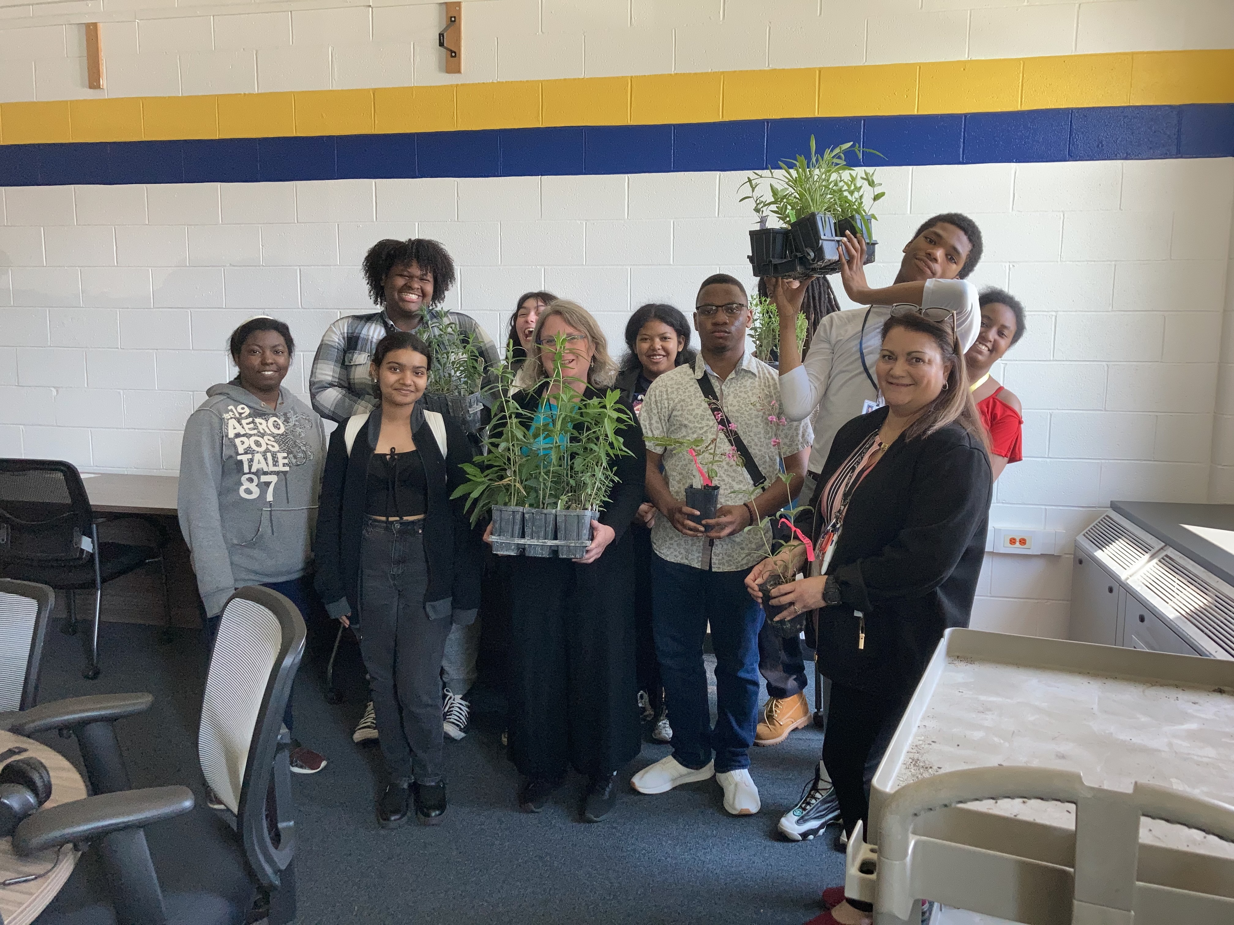 The image shows a group of smiling students standing together in a classroom. They are holding small potted plants