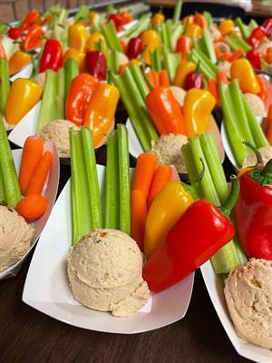 The image shows a close-up view of a platter filled with colorful vegetable sticks and dip. The platter contains celery and carrot sticks cut into thin, elongated pieces and arranged vertically, interspersed with slices of red, orange and yellow bell peppers. In front of the vegetable arrangement are several small round containers, likely filled with various dips or spreads to accompany the fresh, crunchy vegetables. The bright, vibrant colors of the vegetables against the white platter create an appealing and appetizing display.